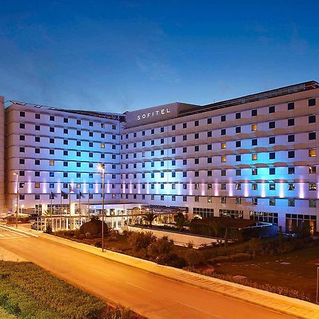 Sofitel Athens Airport Hotel Spata  Bagian luar foto The photo shows a large modern hotel building, likely named "Sofitel," illuminated with blue and purple lights as evening approaches. The structure features multiple floors and a contemporary design, with large windows and a flat facade. In front of 