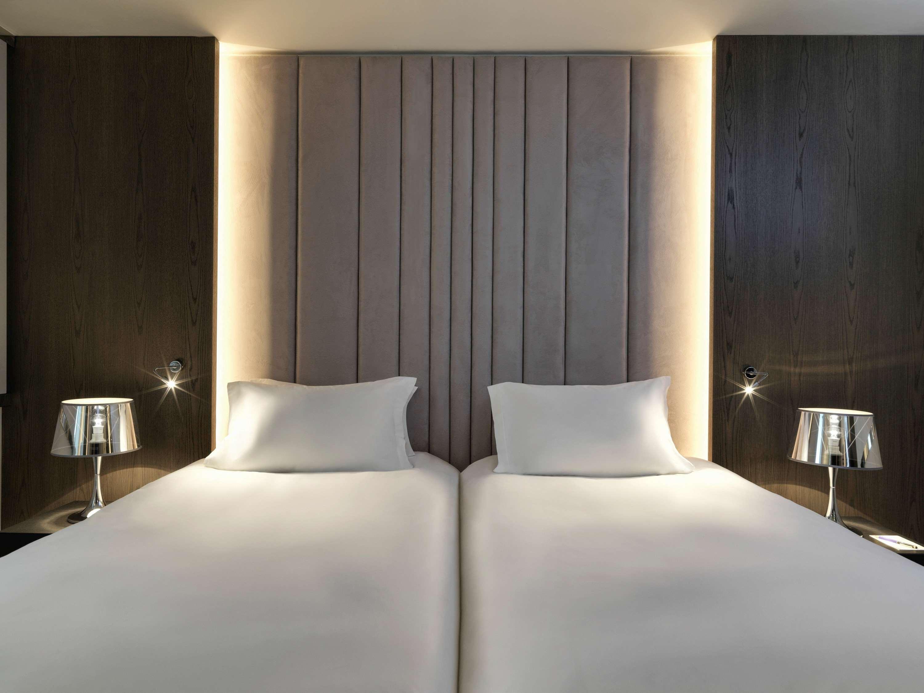 Sofitel Athens Airport Hotel Spata  Bagian luar foto The photo shows a modern bedroom featuring two single beds with white bedding. The beds are positioned against a stylish headboard with vertical panels, illuminated by soft lighting. On either side of the beds, there are sleek, minimalist bedside lam