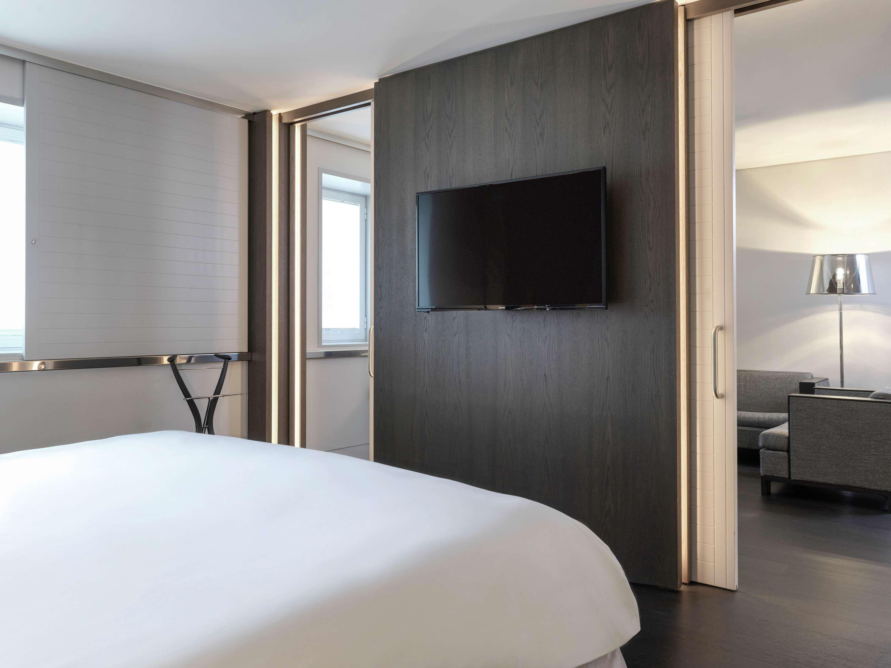 Sofitel Athens Airport Hotel Spata  Bagian luar foto The photo shows a modern hotel room. In the foreground, there is a bed with white linens. Behind the bed, there's a partition wall that includes a flat-screen TV mounted on it. To the sides, there are large windows with light curtains that allow some