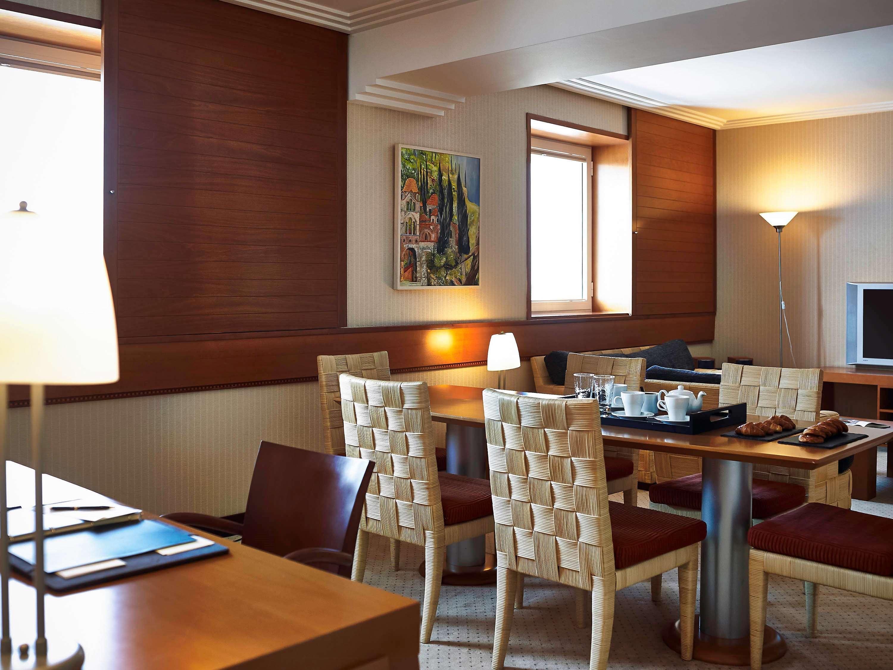 Sofitel Athens Airport Hotel Spata  Bagian luar foto The photo shows a cozy dining area, likely in a hotel or a modern apartment. There is a wooden table surrounded by four chairs with woven backs. On the table, there's a tray with breakfast items, including some pastries. The walls are adorned with wa