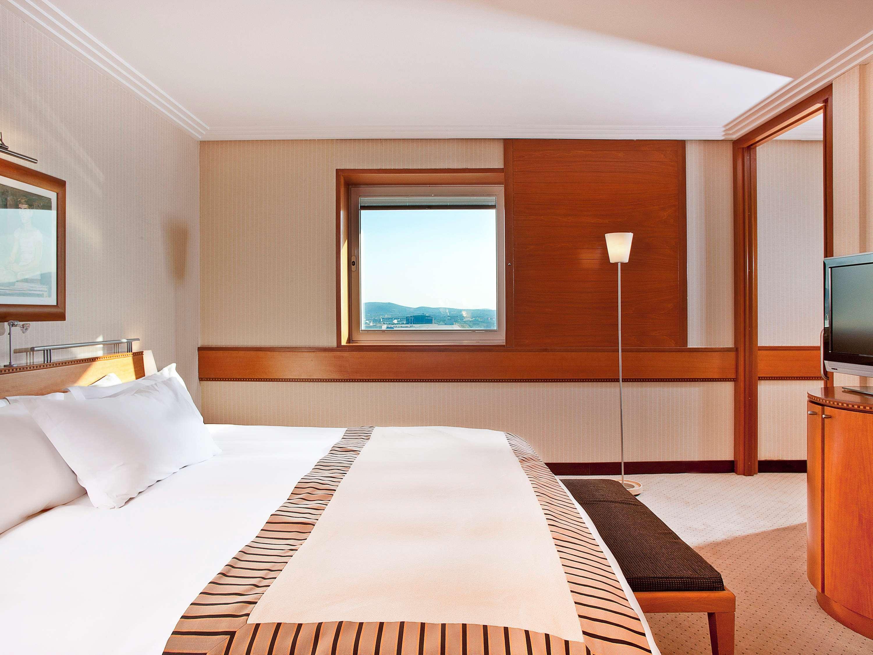 Sofitel Athens Airport Hotel Spata  Bagian luar foto The image shows a stylish hotel room with a modern design. It features a large bed with white linens and a decorative blanket. There is a sitting area with a bench at the foot of the bed. A window provides a view of the outside, allowing natural ligh