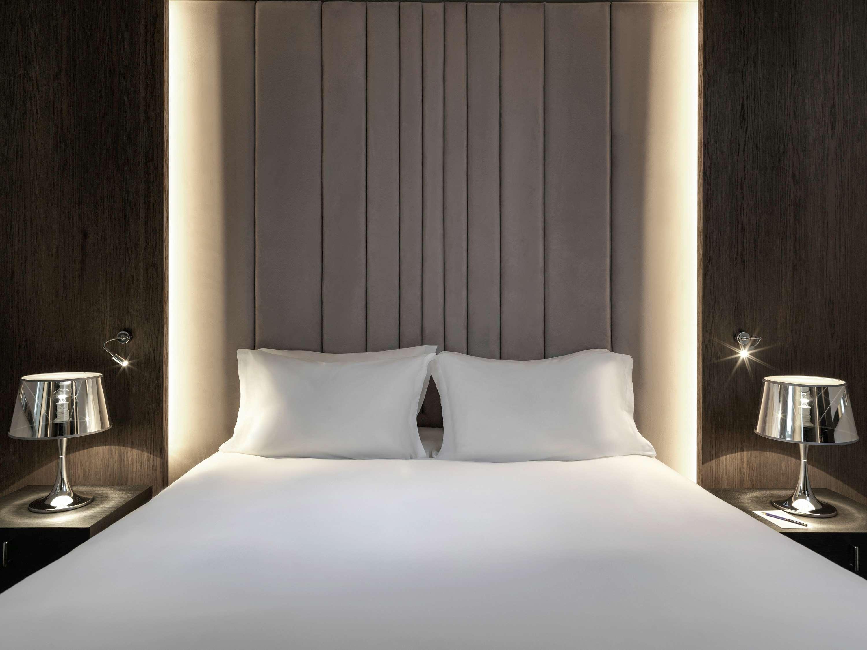 Sofitel Athens Airport Hotel Spata  Bagian luar foto The photo shows a modern bedroom featuring a double bed with a simple yet elegant design. The headboard is upholstered in a light fabric with vertical paneling, enhanced by soft lighting around it. On either side of the bed, there are stylish bedside