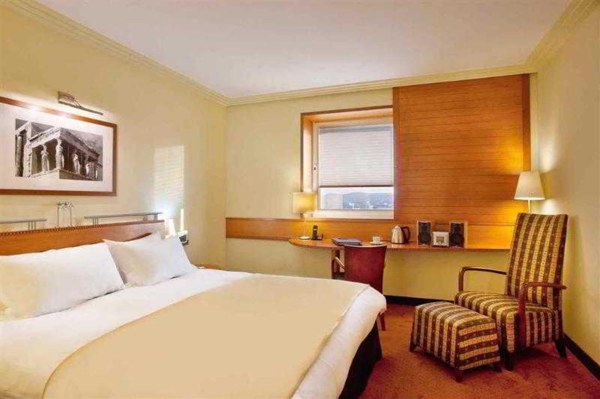 Sofitel Athens Airport Hotel Spata  Bagian luar foto The photo depicts the interior of a hotel room. It features a large bed with white linens and a beige blanket. There is a window with blinds, allowing some natural light to enter the space. Beside the bed, there is a bedside lamp and a piece of artwo