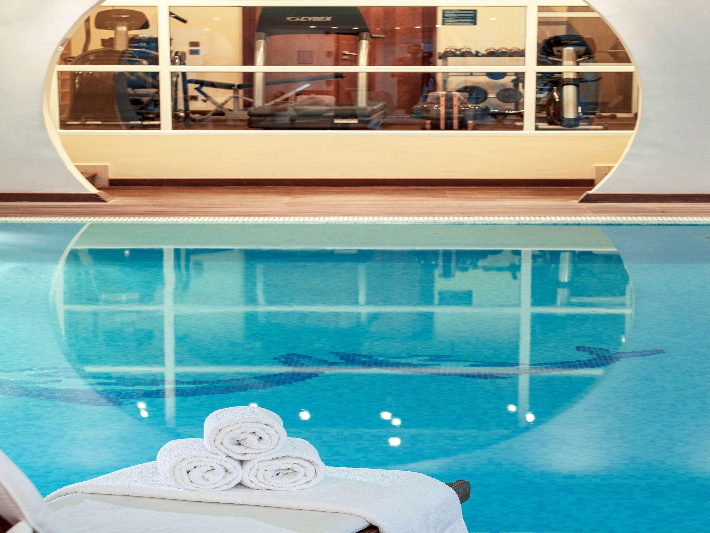Sofitel Athens Airport Hotel Spata  Bagian luar foto The photo shows a tranquil indoor swimming pool area. The water is clear and blue, and there are neatly stacked white towels on a lounge chair. In the background, there is a glimpse of a fitness area visible through large glass windows, featuring var