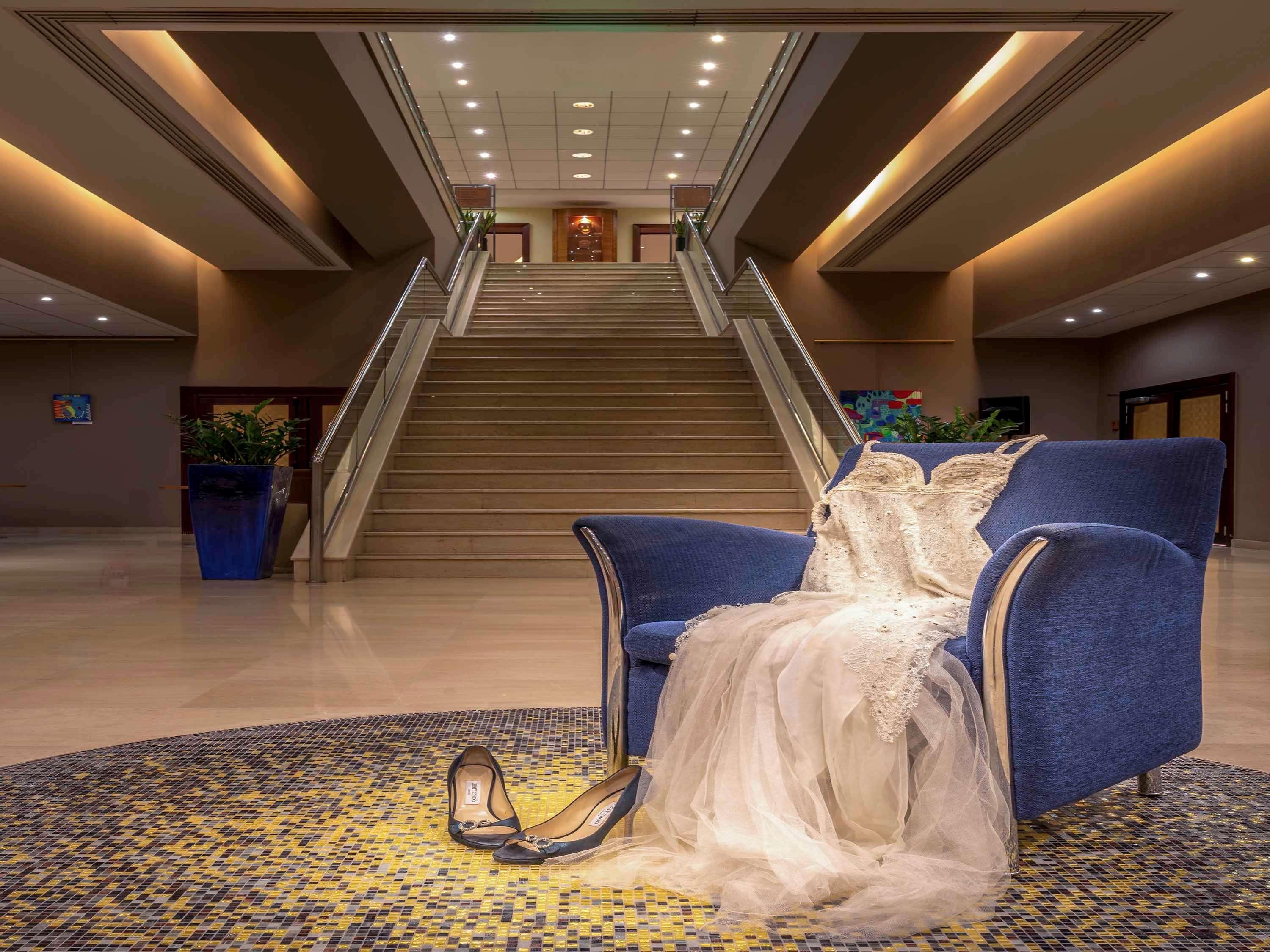 Sofitel Athens Airport Hotel Spata  Bagian luar foto The photo depicts a stylish blue armchair positioned in a spacious lobby area. On the chair, there is a wedding dress draped elegantly, and nearby, a pair of wedding shoes is placed on the floor. The background features a grand staircase leading up, 