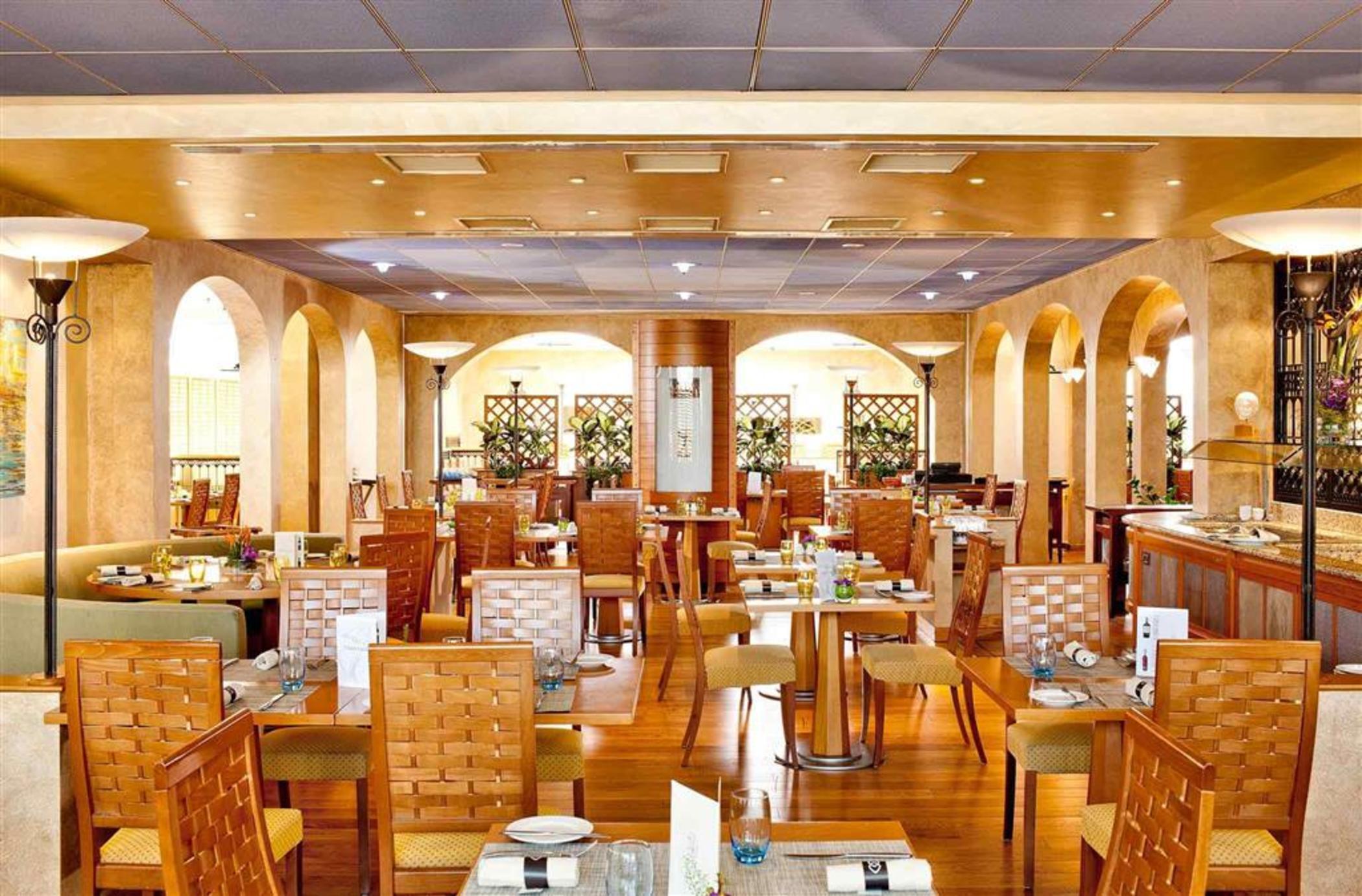 Sofitel Athens Airport Hotel Spata  Bagian luar foto The photo shows the interior of a restaurant. The space features warm, golden tones with wooden furniture. There are several tables with chairs arranged throughout the room, and the tables appear to be set for dining with some plates and utensils. Ar