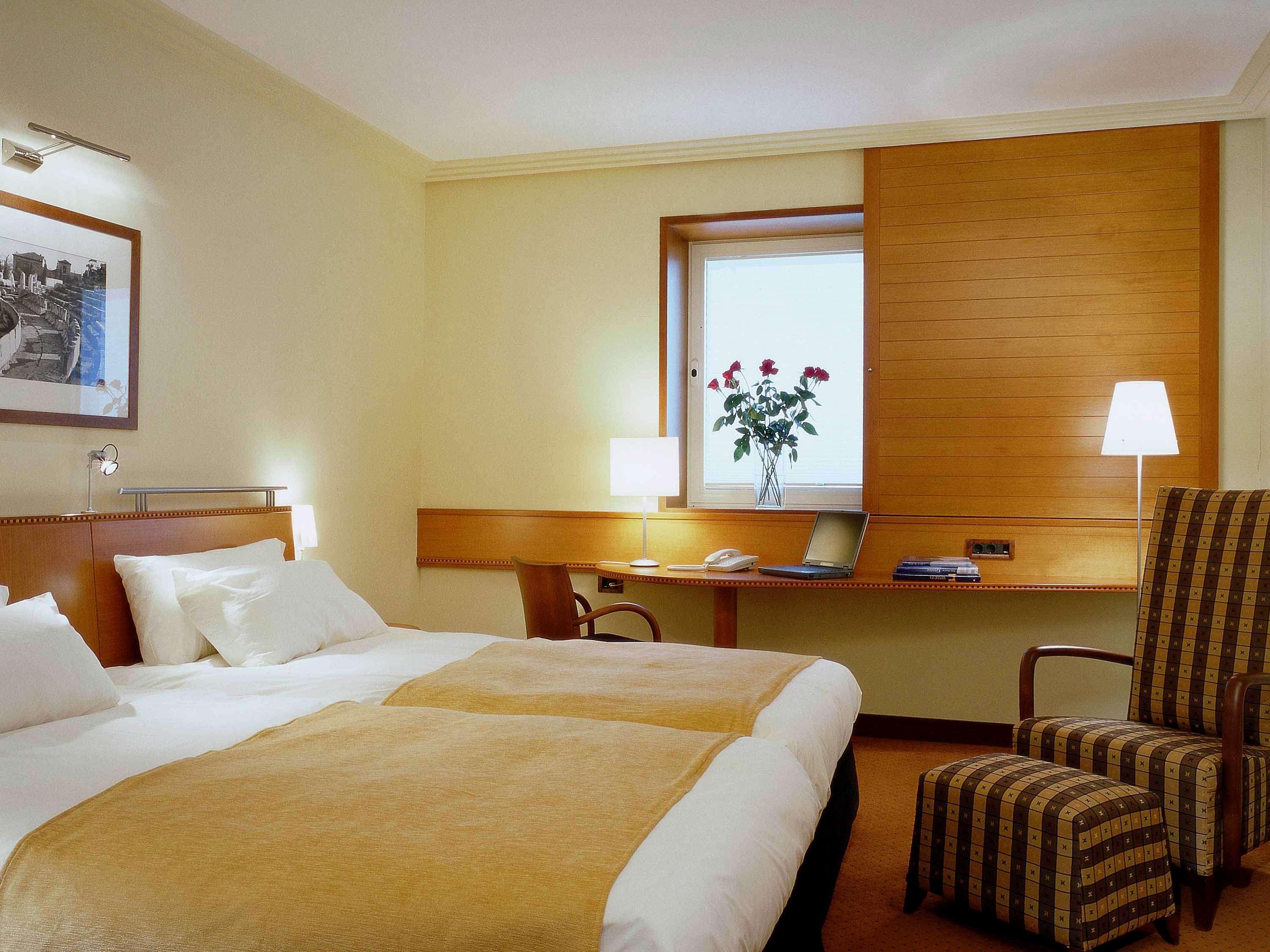 Sofitel Athens Airport Hotel Spata  Bagian luar foto The photo shows a cozy hotel room. It features two twin beds with light-colored bedding and taupe blankets. There is a wooden headboard above the beds. A window with light drapes allows natural light to enter, and there are decorative roses on the wi