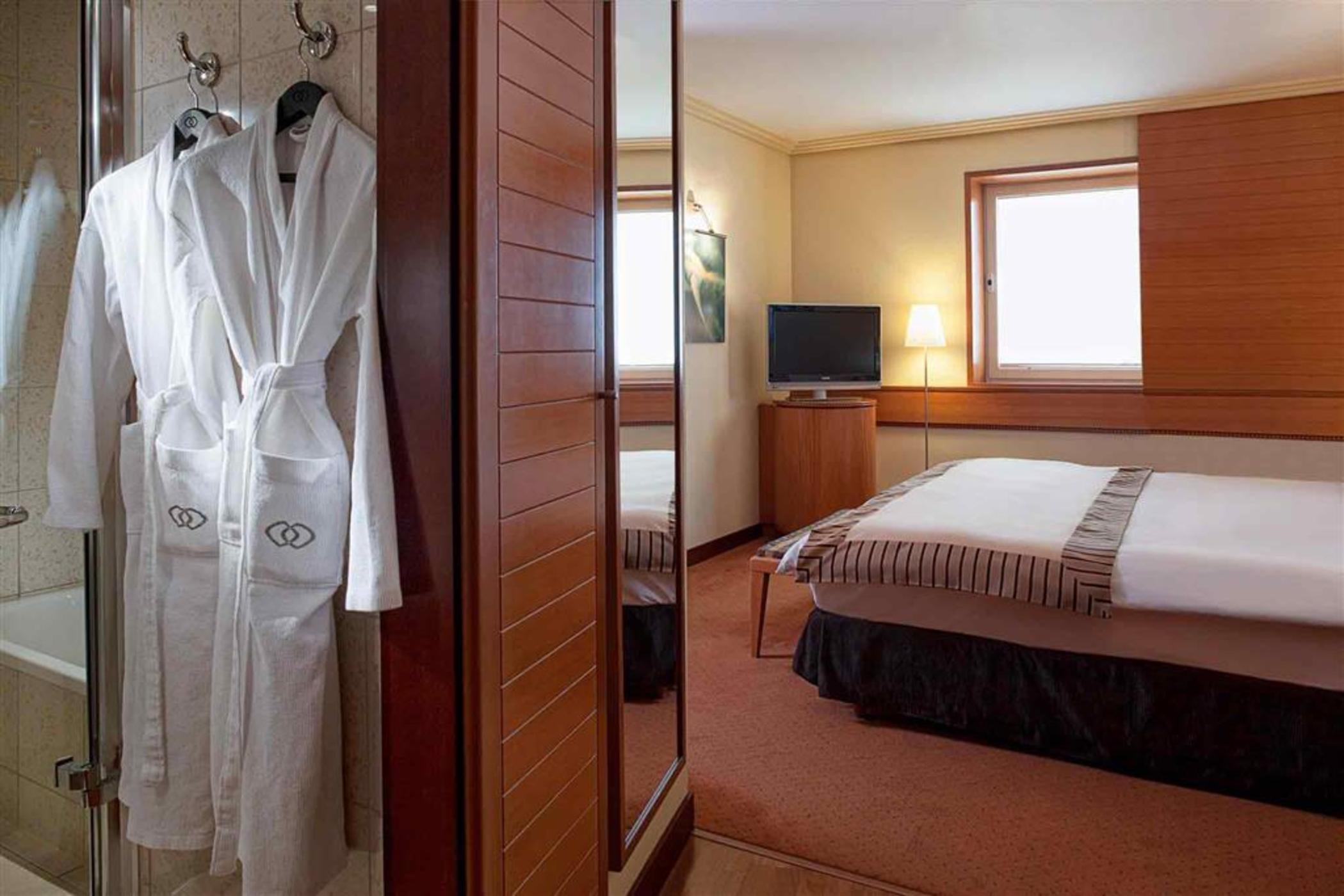 Sofitel Athens Airport Hotel Spata  Bagian luar foto The photo depicts a cozy hotel room. On one side, there's a set of wooden wardrobes, and hanging from one of them is a white bathrobe. The room features a neatly made bed with a striped blanket and decorative pillows. There's a small television mount