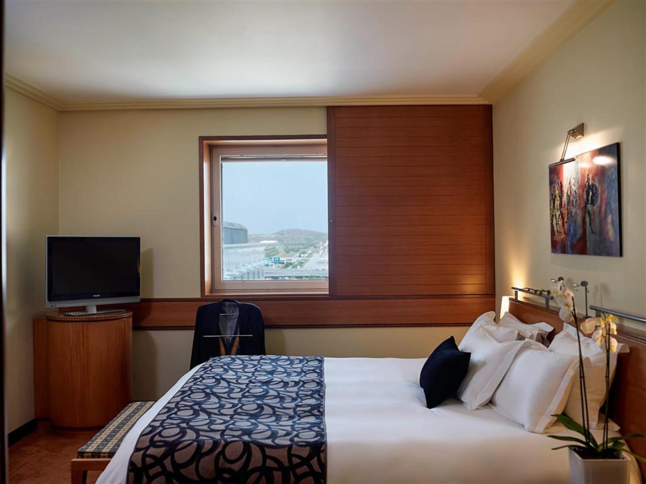Sofitel Athens Airport Hotel Spata  Bagian luar foto The photo depicts a cozy hotel room featuring a large bed with white linens and a patterned blue throw. There are several pillows on the bed, and a bedside table with a small lamp. A window allows natural light to enter the room, and it has wooden bl