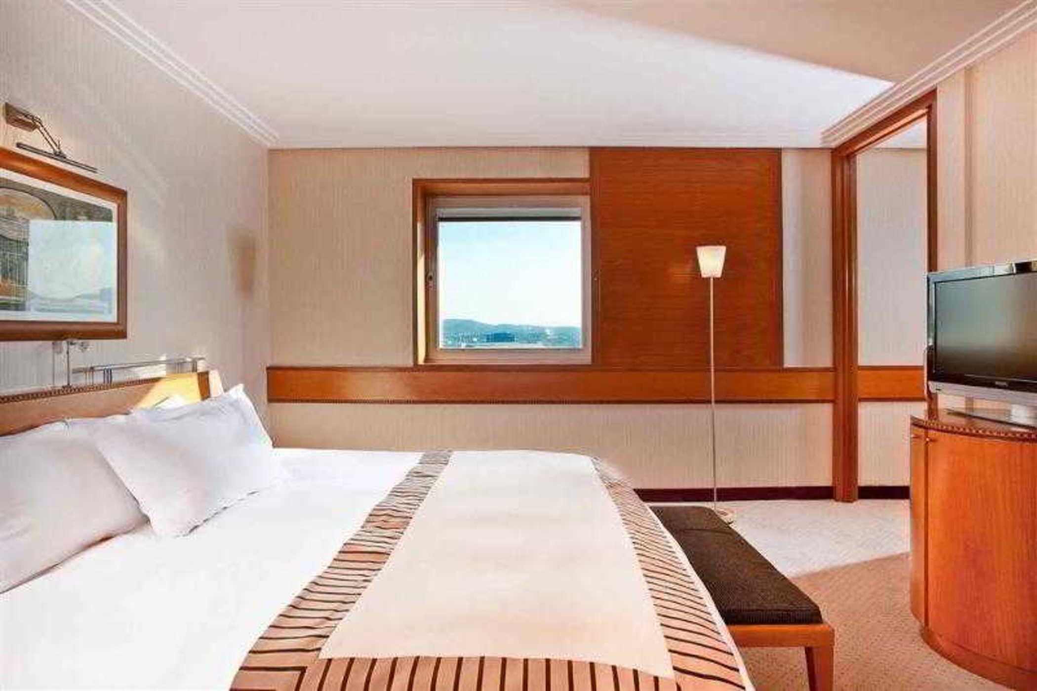 Sofitel Athens Airport Hotel Spata  Interior foto The photo shows a modern hotel room featuring a large bed with white linens and a decorative blanket. There's a wooden bench at the foot of the bed. A window with a view is visible, allowing natural light to enter the room. On one side, there's a lam