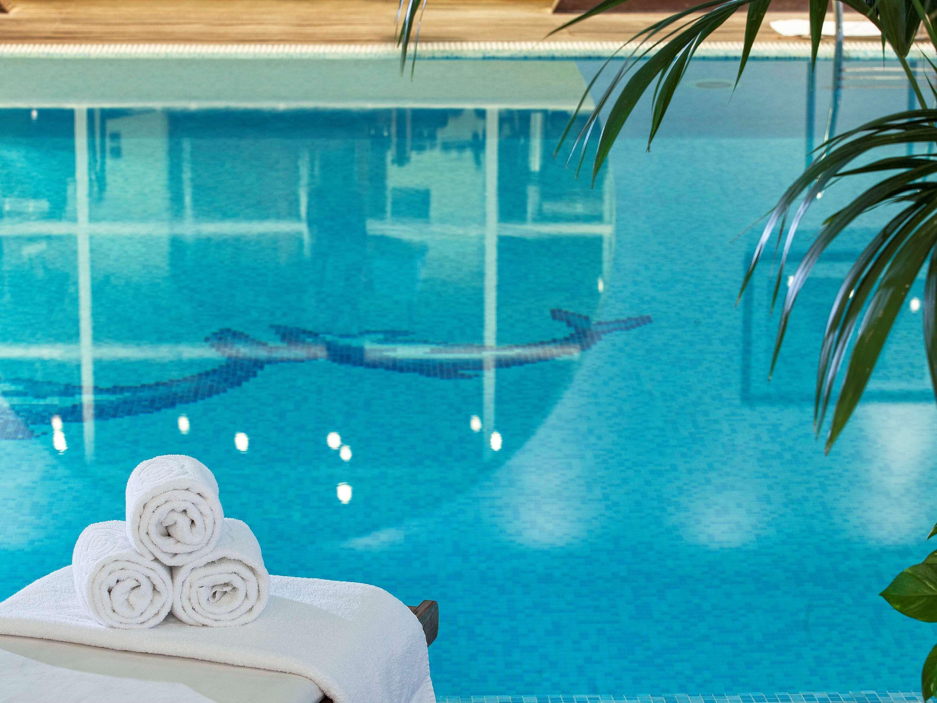Sofitel Athens Airport Hotel Spata  Bagian luar foto The photo shows a serene swimming pool with clear, turquoise water. In the foreground, there are neatly rolled towels placed on a lounge chair. The water reflects soft lighting and has a tiled pattern visible beneath the surface, while there are hint