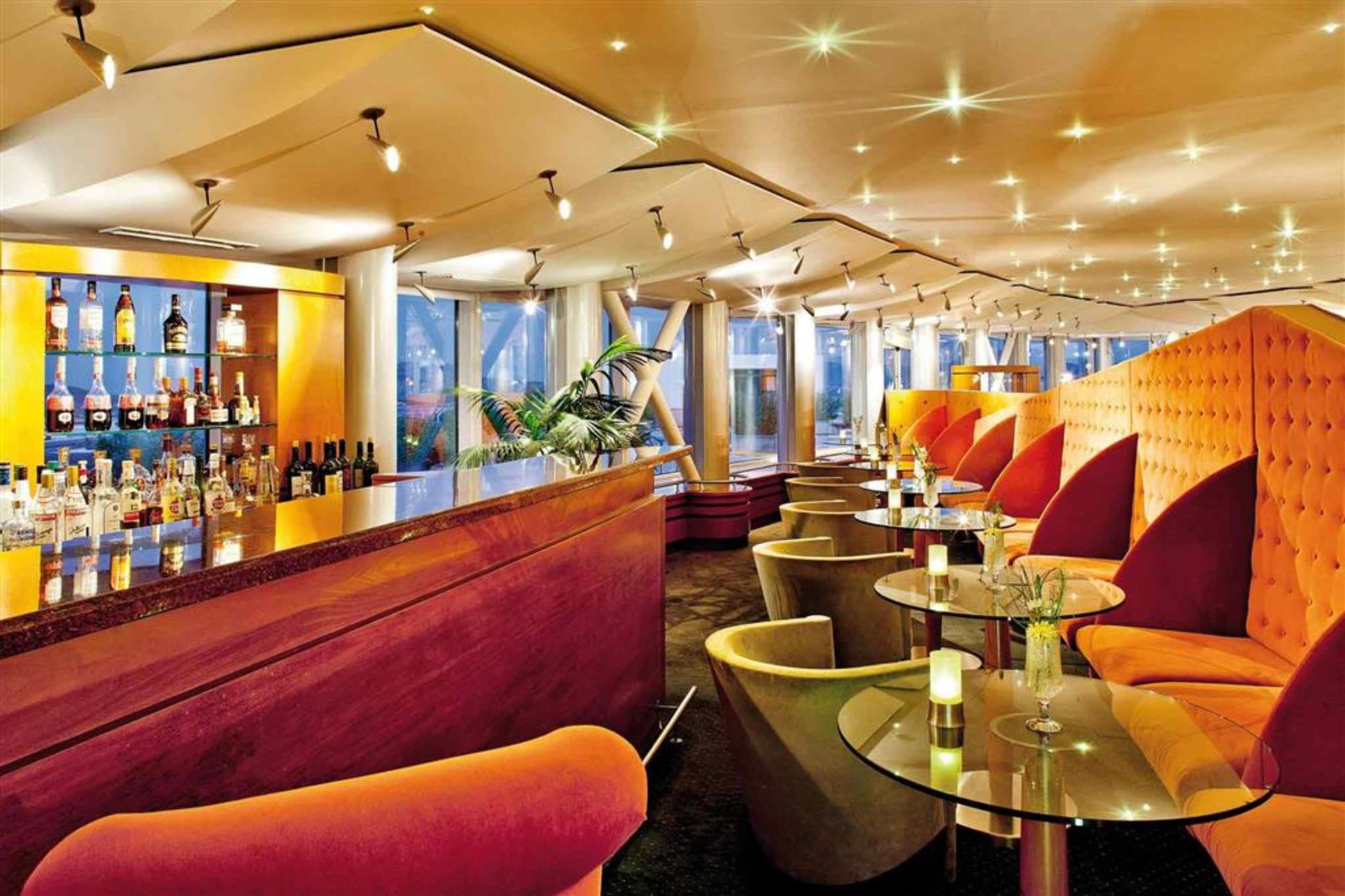 Sofitel Athens Airport Hotel Spata  Bagian luar foto The photo shows a stylish bar interior featuring a modern design. There's a long bar counter with an array of bottles displayed on shelves behind it. The seating area includes cozy, brightly colored chairs and small tables. The lighting is soft and h