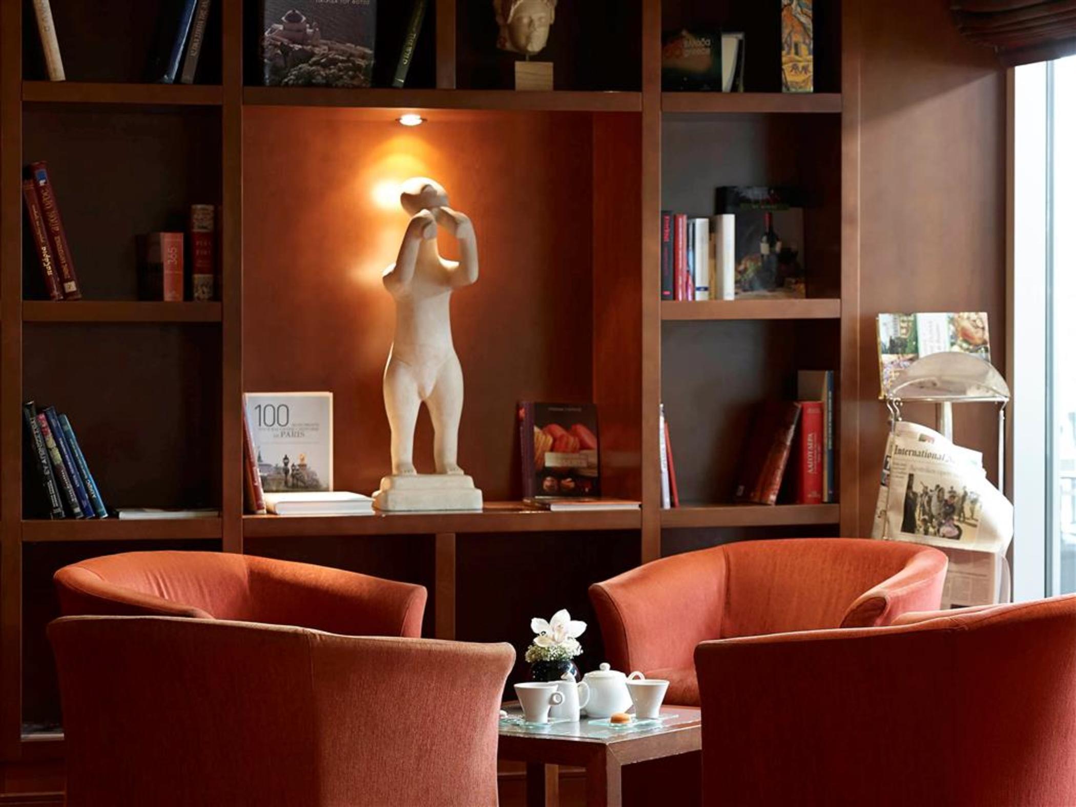 Sofitel Athens Airport Hotel Spata  Bagian luar foto The photo shows a cozy interior space, likely a lounge or reading area. In the center, there are two orange chairs facing each other, around a small table. On the table, there appears to be a teapot and teacups, along with a small flower arrangement.