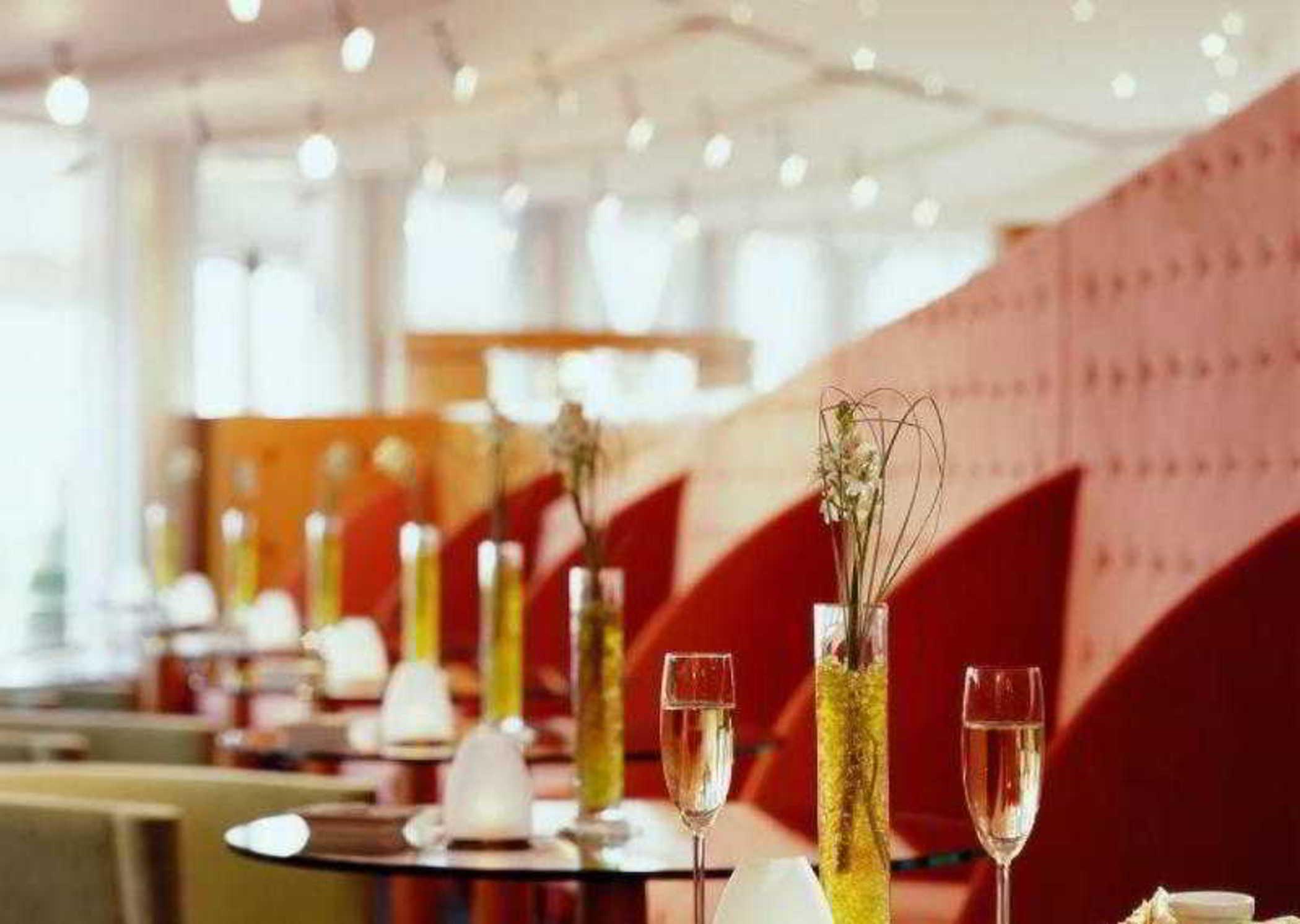 Sofitel Athens Airport Hotel Spata  Bagian luar foto The image depicts the interior of a modern restaurant or lounge. The space features elegantly arranged tables with small vases of greenery as centerpieces. Each table has stemmed glasses, possibly for champagne or cocktails. The decor includes soft, 