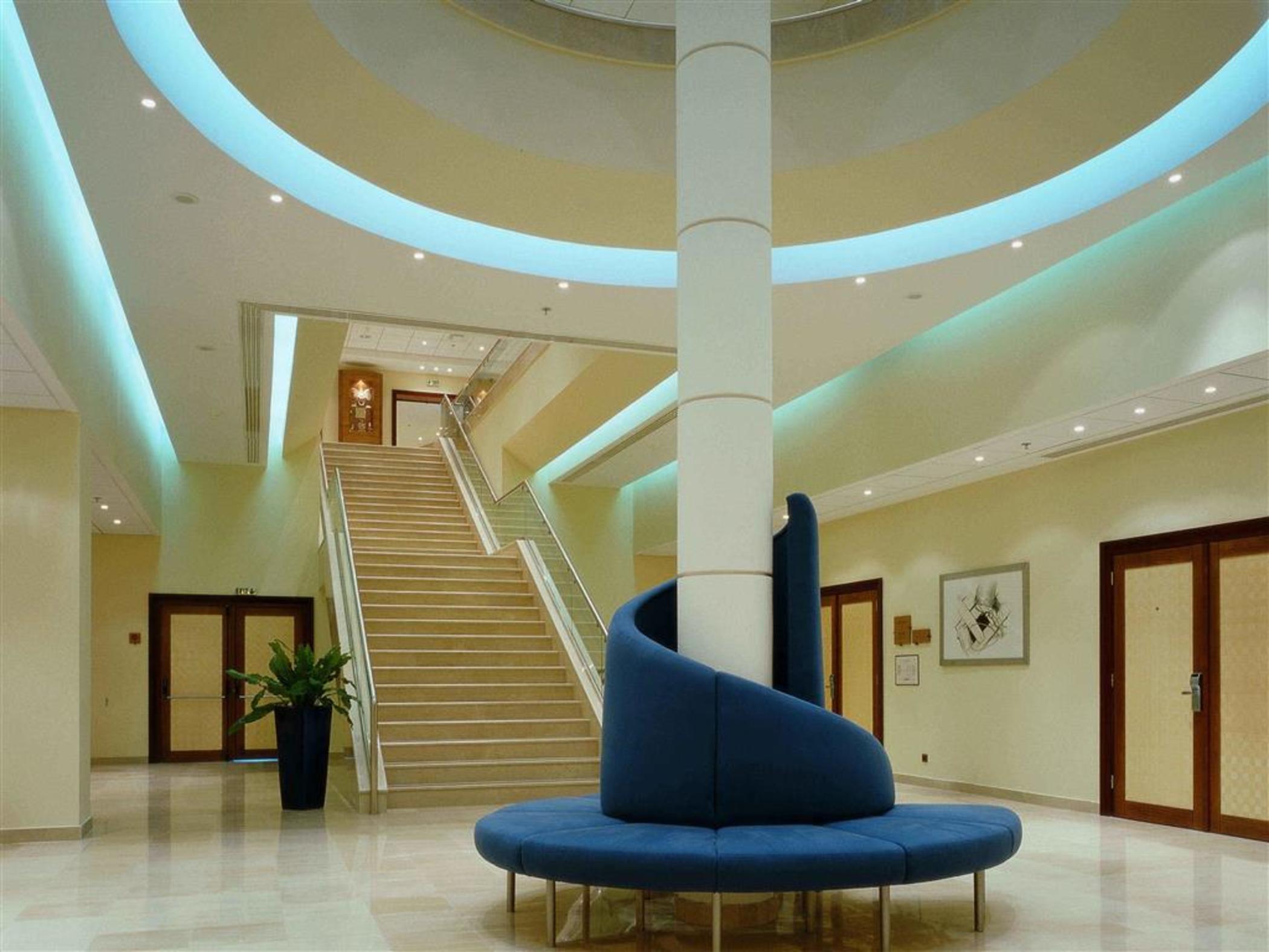 Sofitel Athens Airport Hotel Spata  Interior foto The photo shows a modern lobby area with an elegant design. There is a large, spiraled seating fixture in blue, which is situated in the center. Surrounding this seating area, the floor is made of polished tiles that reflect light. 

Behind the seati