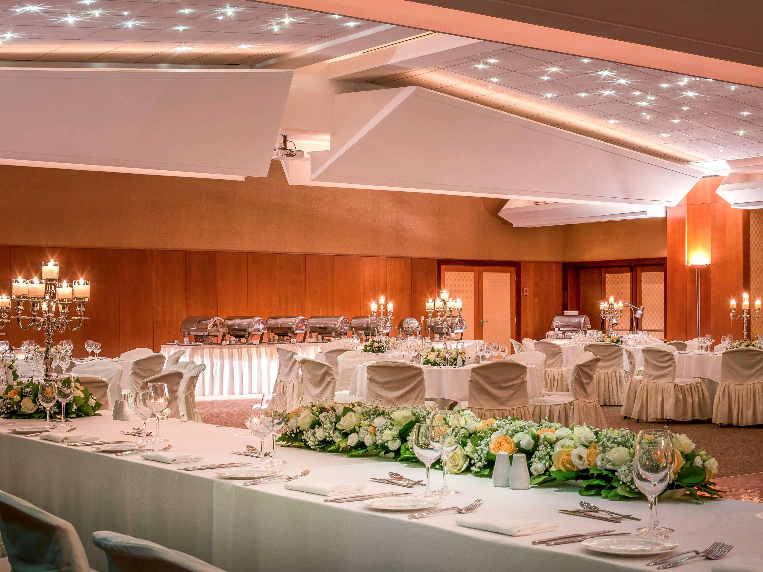 Sofitel Athens Airport Hotel Spata  Bagian luar foto The photo shows an elegantly decorated banquet or event hall. The room features a large table set with fine dining ware, including crystal glasses and silver utensils. There are multiple round tables arranged around the space, each adorned with white