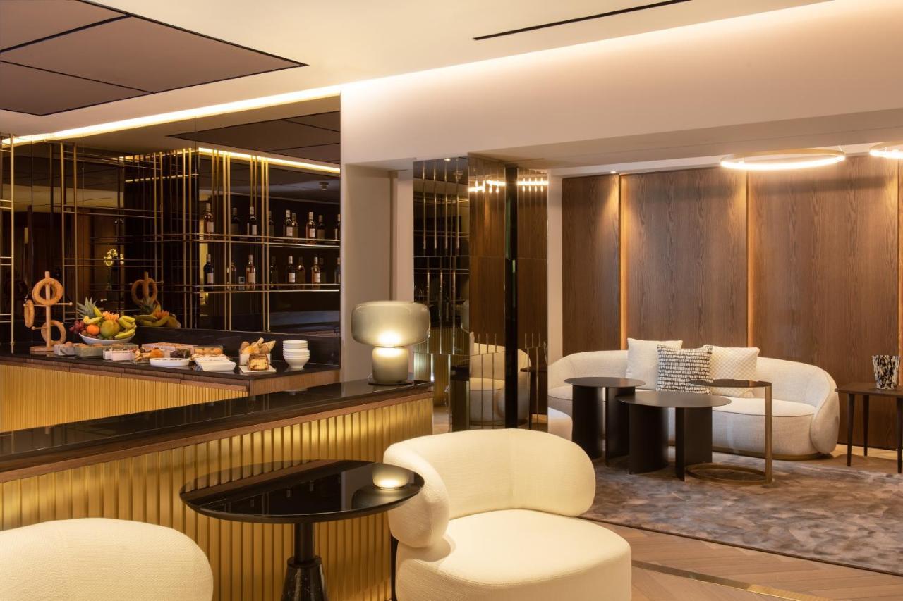Sofitel Athens Airport Hotel Spata  Bagian luar foto The photo depicts a modern lounge or bar interior. It features a counter with a sleek design that has golden accents. Behind the counter, there are shelves displaying various bottles. The seating area includes two stylish, round chairs and a small bl