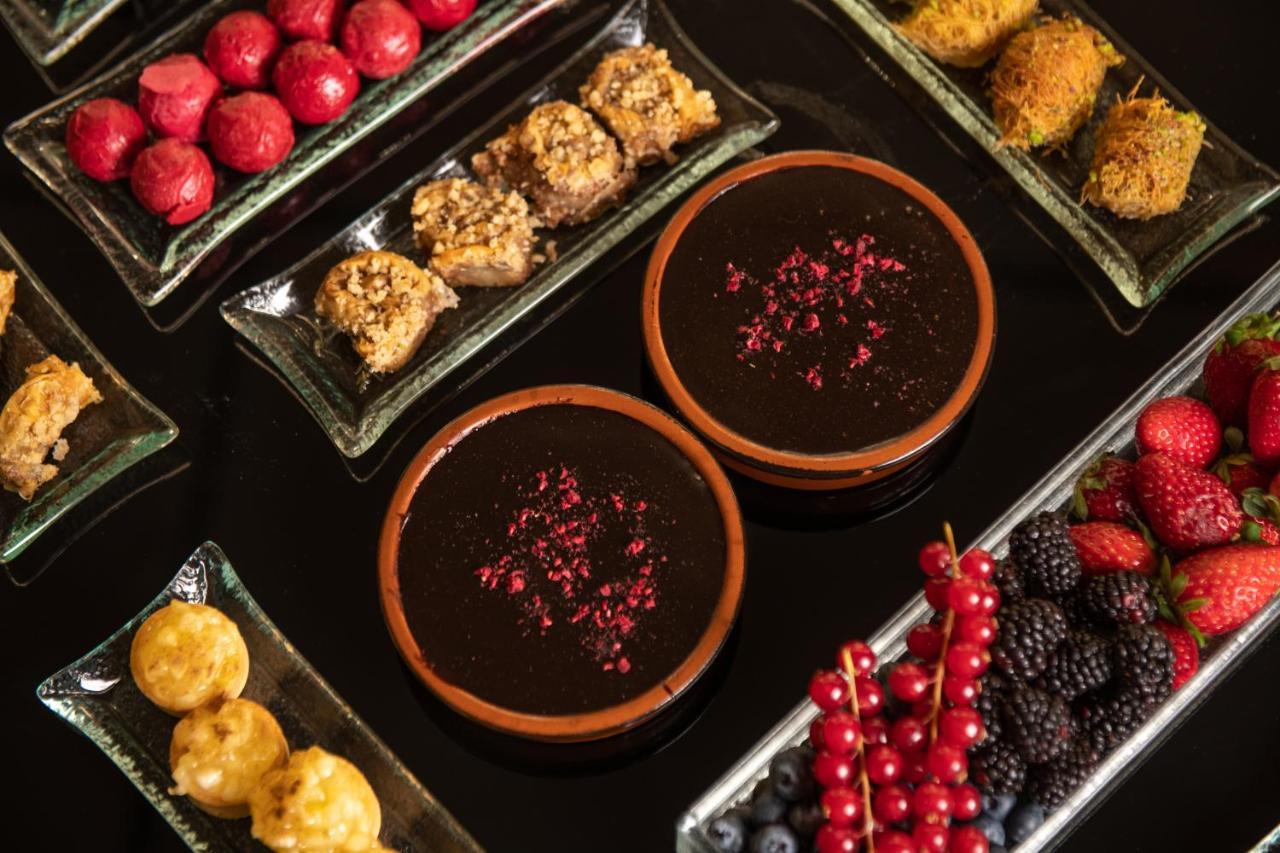 Sofitel Athens Airport Hotel Spata  Bagian luar foto The image shows a beautifully arranged assortment of desserts and sweets on a dark surface. There are two brown bowls filled with a thick, dark dessert, garnished with rose petals. Surrounding the bowls are various plates containing a variety of trea