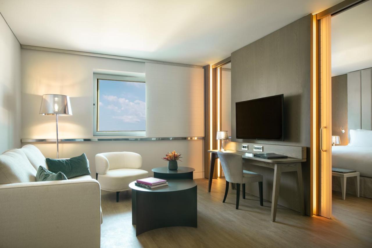 Sofitel Athens Airport Hotel Spata  Bagian luar foto The photo displays a modern hotel room or suite. There is a small seating area with a white armchair and a light gray sofa. A round coffee table sits in front of the seating area. A small desk and chair are positioned against one wall, with a televis