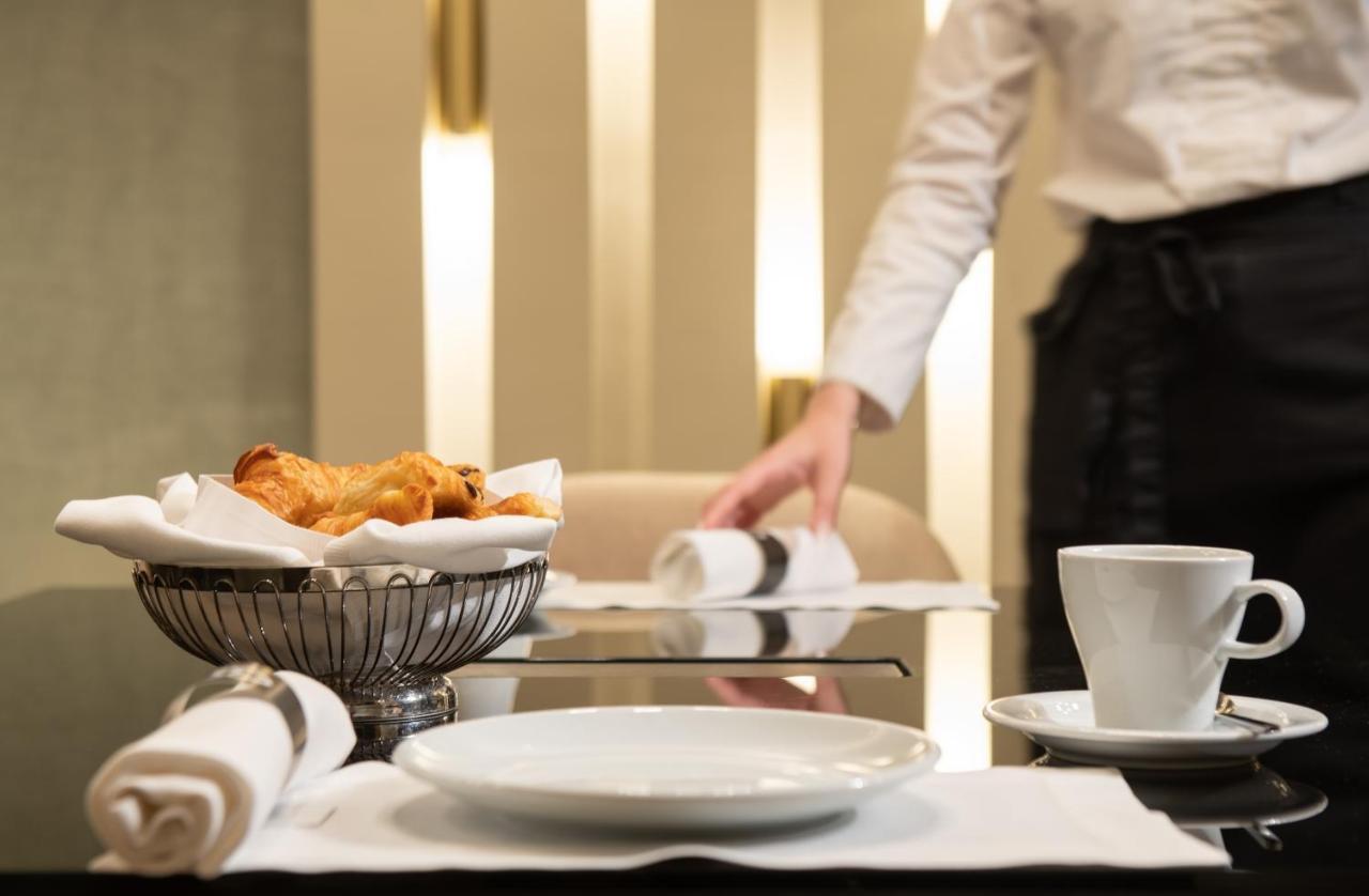 Sofitel Athens Airport Hotel Spata  Bagian luar foto The photo shows a dining table setting in a restaurant. On the table, there is a white plate, a small cup, and a silver bowl containing pastries, likely croissants. The surrounding area has a clean, modern aesthetic with soft lighting. In the backgro