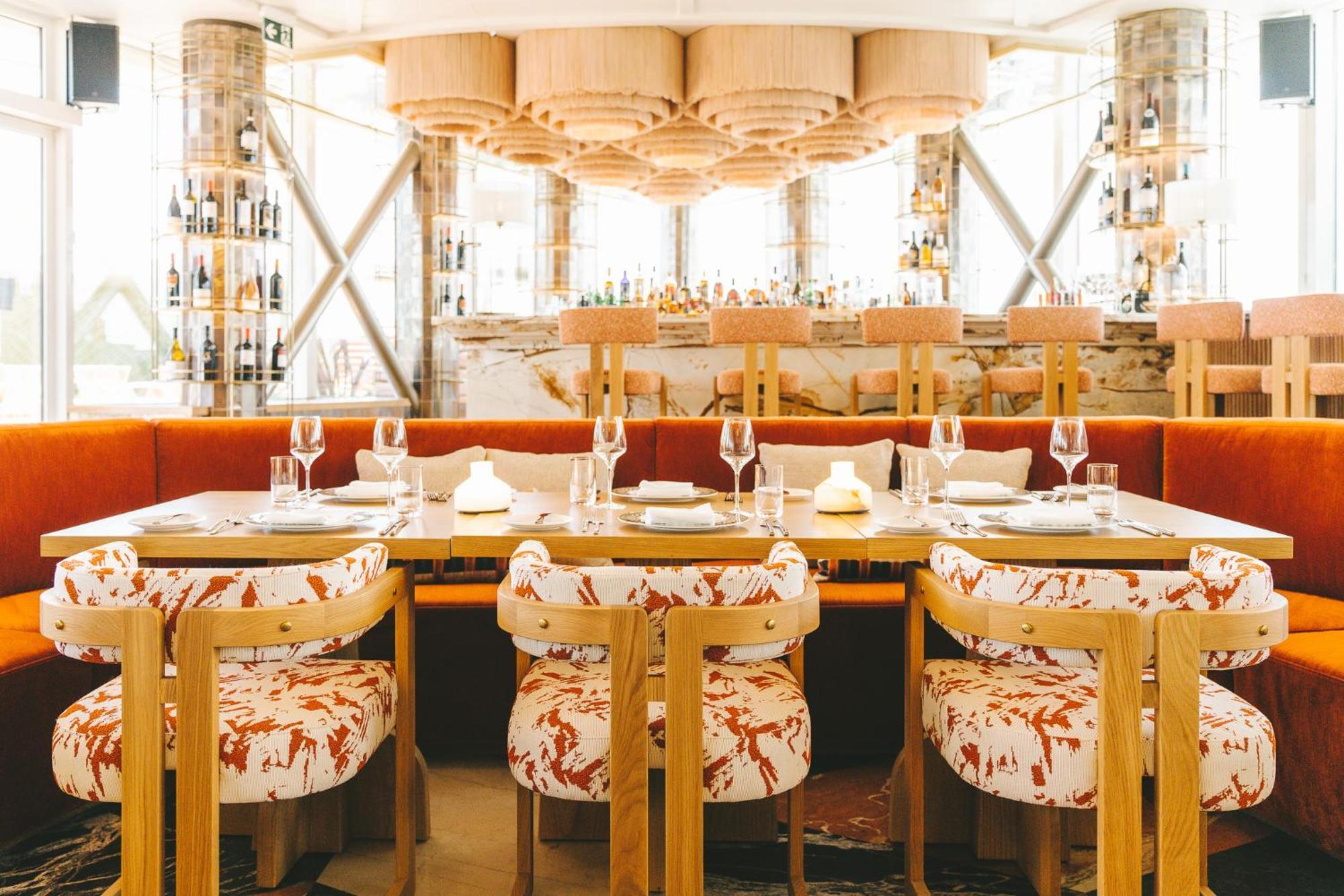 Sofitel Athens Airport Hotel Spata  Bagian luar foto The photo depicts a stylish restaurant interior. It features a modern design with a warm color palette. In the foreground, there are several dining tables surrounded by unique chairs that have a vibrant floral pattern. The chairs are made of wood wit