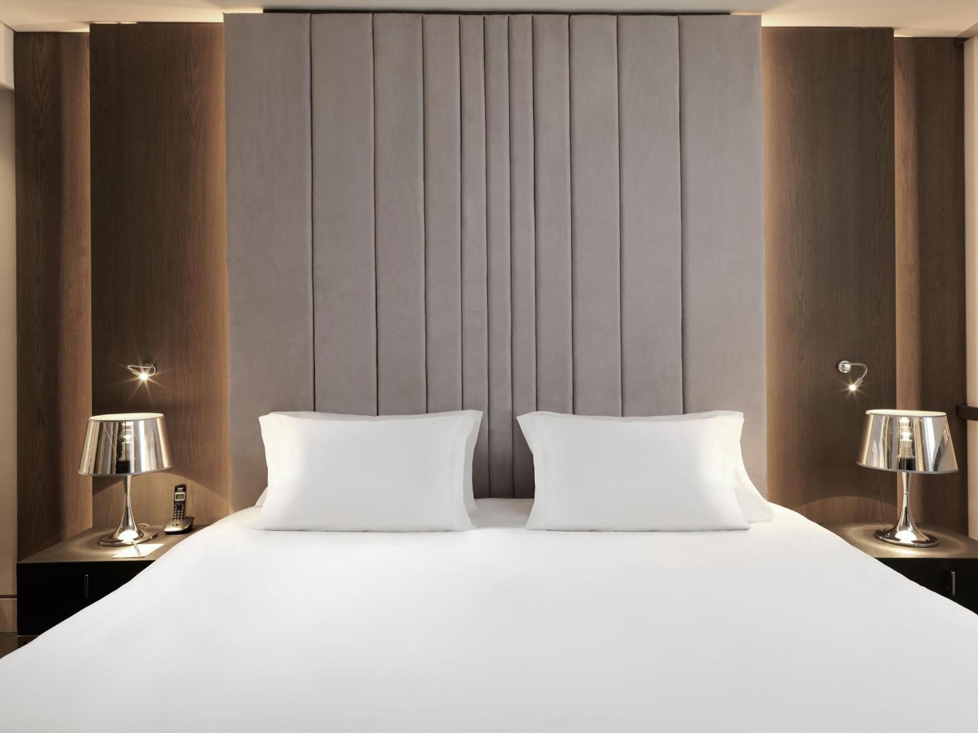 Sofitel Athens Airport Hotel Spata  Bagian luar foto The image shows a modern bedroom featuring a large bed with a minimalistic design. The bed has two white pillows and is placed against a stylish padded headboard that has vertical lines. On either side of the bed, there are sleek bedside tables, each