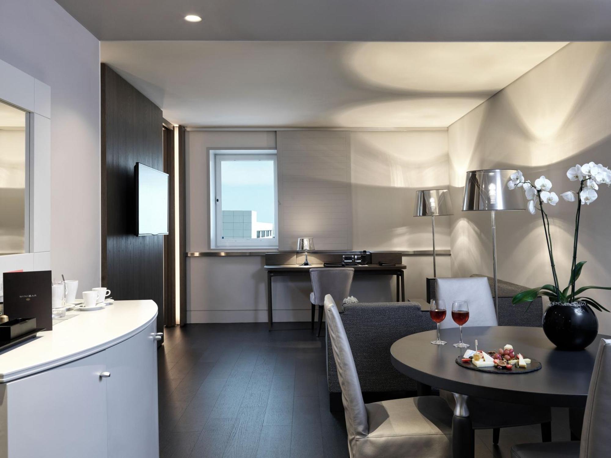 Sofitel Athens Airport Hotel Spata  Bagian luar foto The photo shows a modern and elegantly furnished living space, likely a hotel room or a suite. The room features:

- A stylish seating area with a gray armchair and a small table.
- A round dining table set for two, with plates of food and drinks.
- 