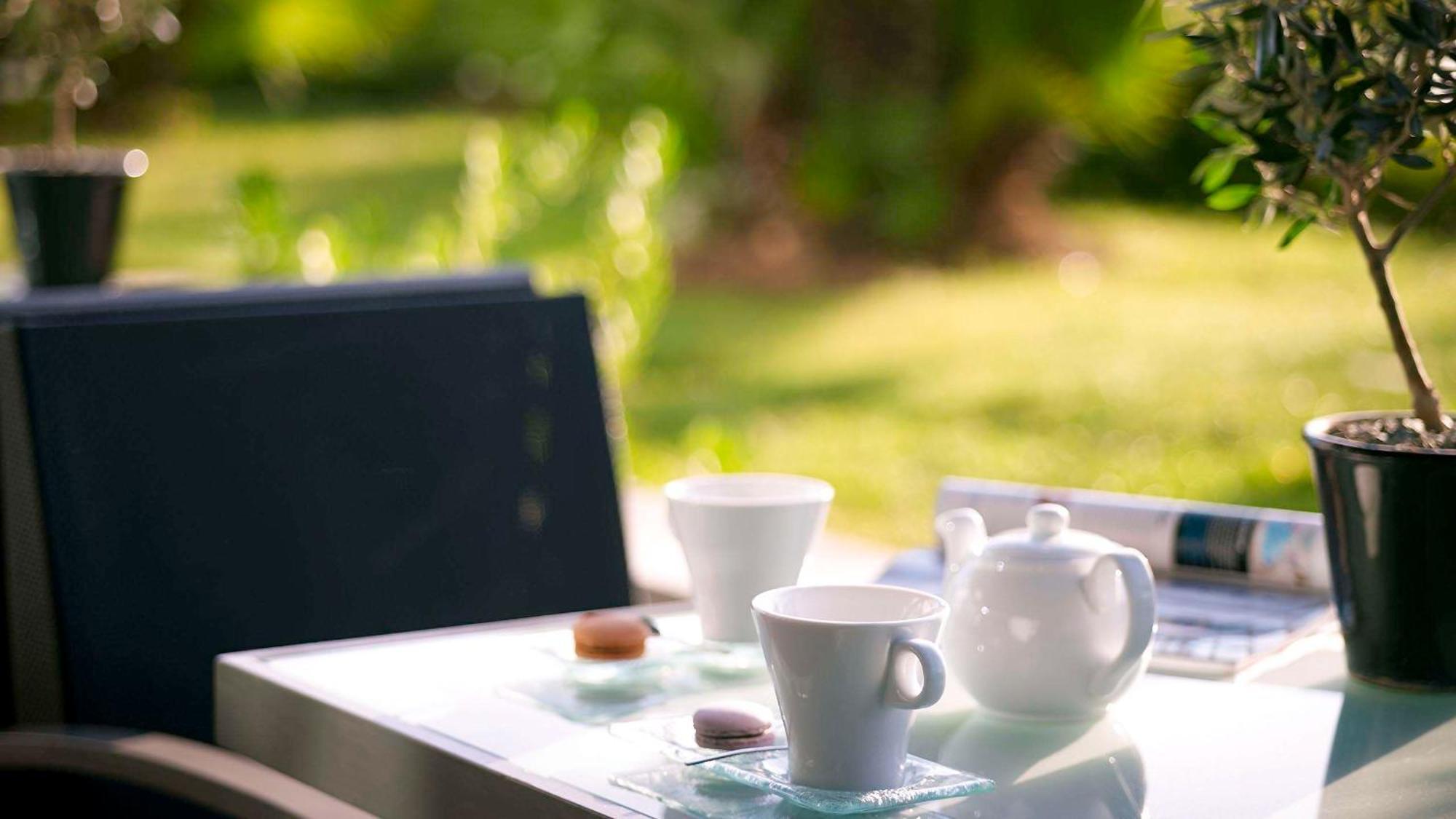 Sofitel Athens Airport Hotel Spata  Bagian luar foto The photo shows a serene outdoor setting with a table featuring a tea setup. There are two white cups, a white teapot, and a small cookie or biscuit placed on the table. The background features lush greenery, suggesting a tranquil garden or patio atm