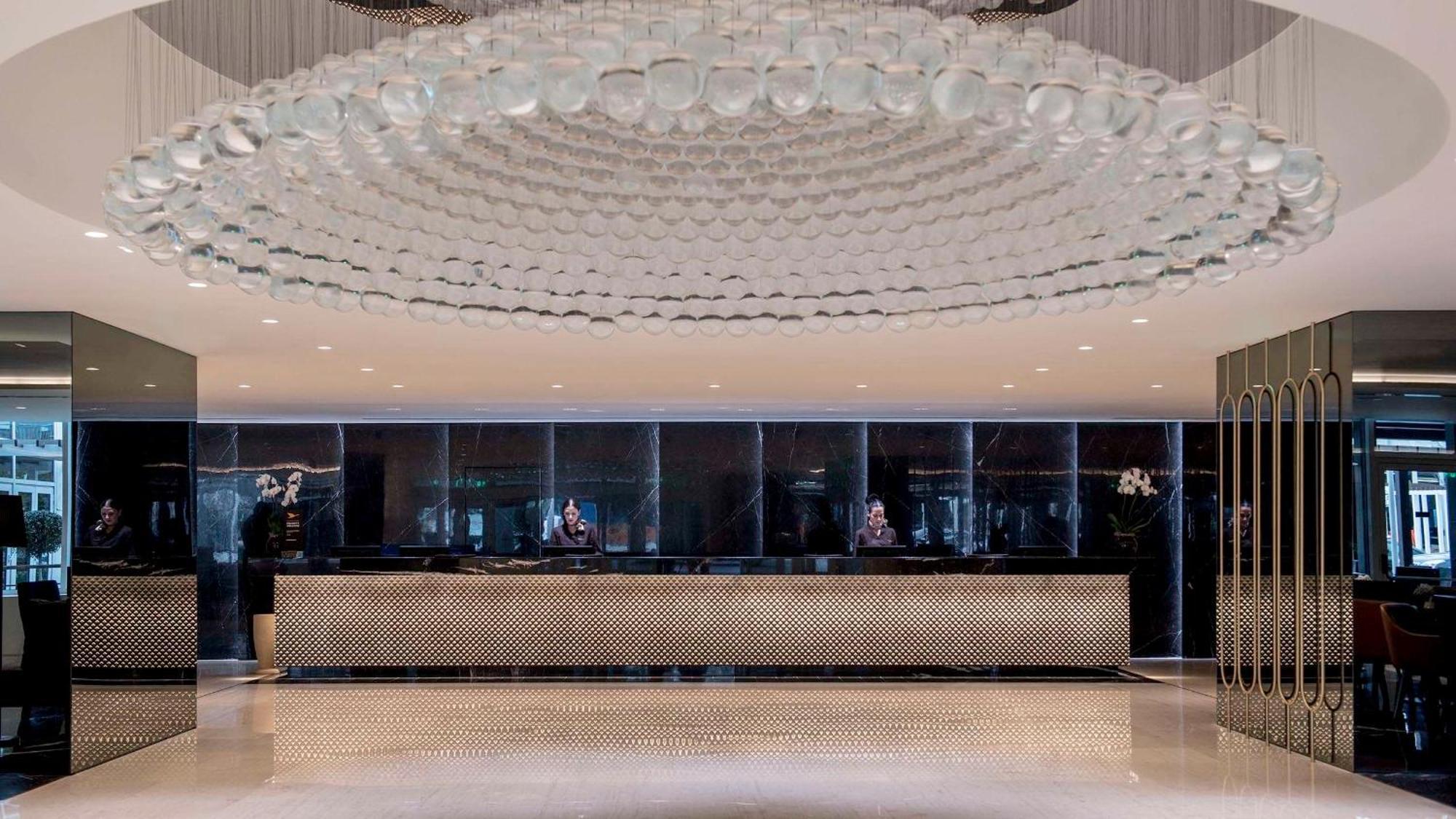 Sofitel Athens Airport Hotel Spata  Bagian luar foto The photo shows a modern hotel lobby. The space features a sleek design with a large, artistic chandelier made of glass or crystal that hangs from the ceiling. The reception desk is centrally located and designed with a polished, reflective surface. 