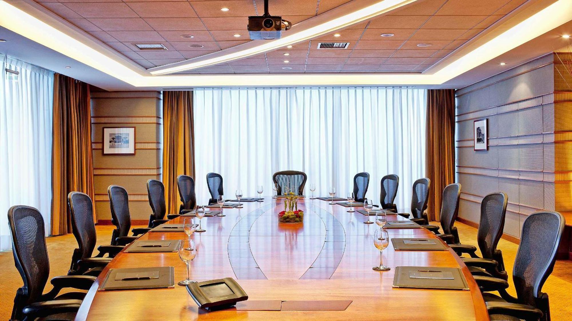 Sofitel Athens Airport Hotel Spata  Bagian luar foto The photo depicts a modern conference room featuring a large, oval wooden table surrounded by several ergonomic chairs. The room has dim lighting and is illuminated by natural light coming in through large windows covered by sheer curtains. There are