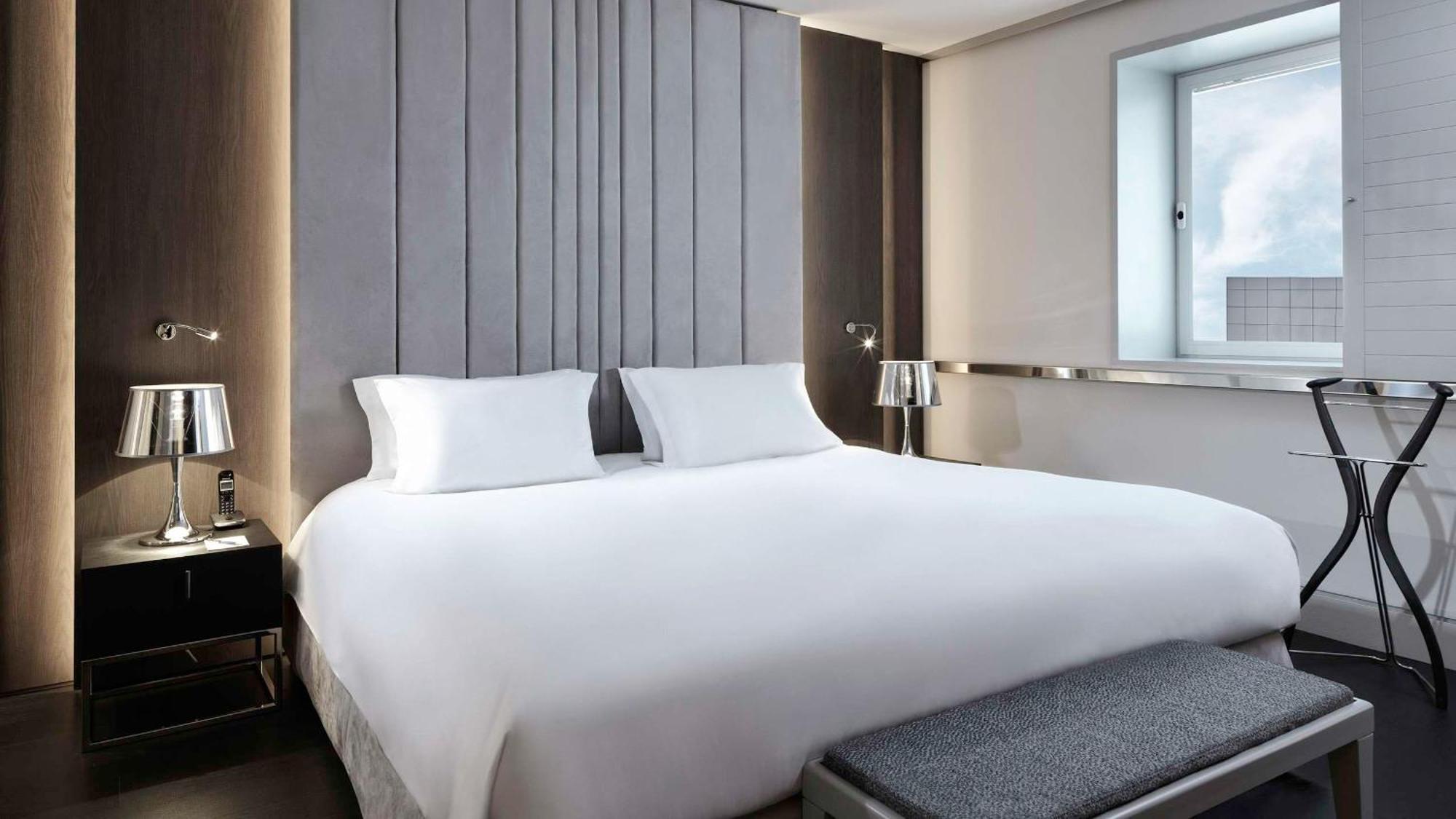 Sofitel Athens Airport Hotel Spata  Bagian luar foto The photo shows a modern hotel room featuring a large bed with white linens and two white pillows. The headboard has a sleek, vertical design, adding a contemporary touch to the room. On either side of the bed, there are stylish bedside tables with t