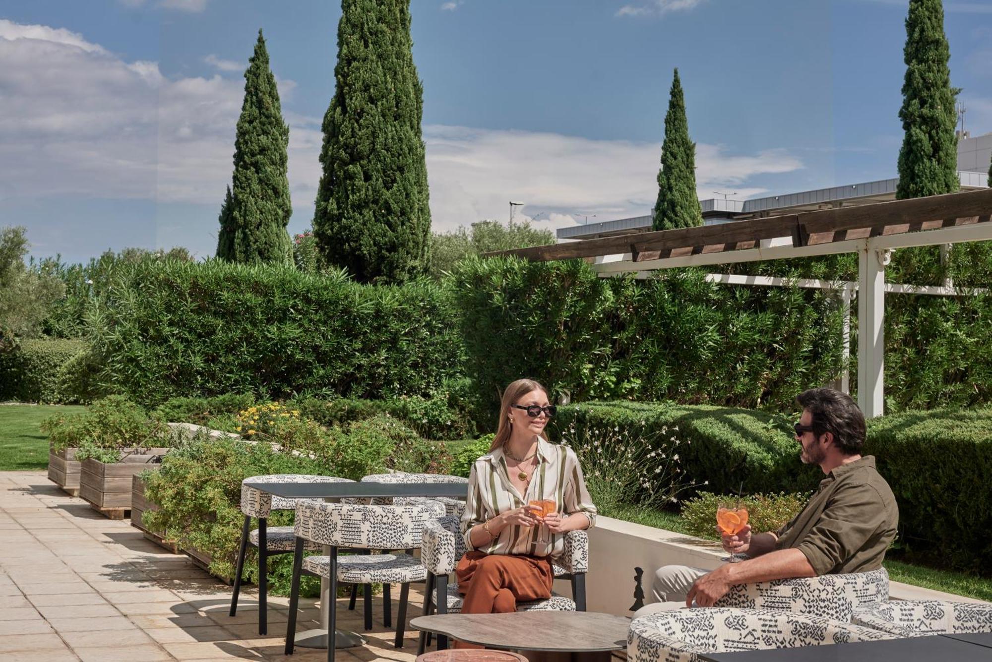 Sofitel Athens Airport Hotel Spata  Bagian luar foto The photo shows two people seated at an outdoor setting, enjoying drinks. They are positioned at a table with decorative tablecloths, surrounded by lush greenery and tall cypress trees. The atmosphere appears relaxed and sunny, suggesting a pleasant 