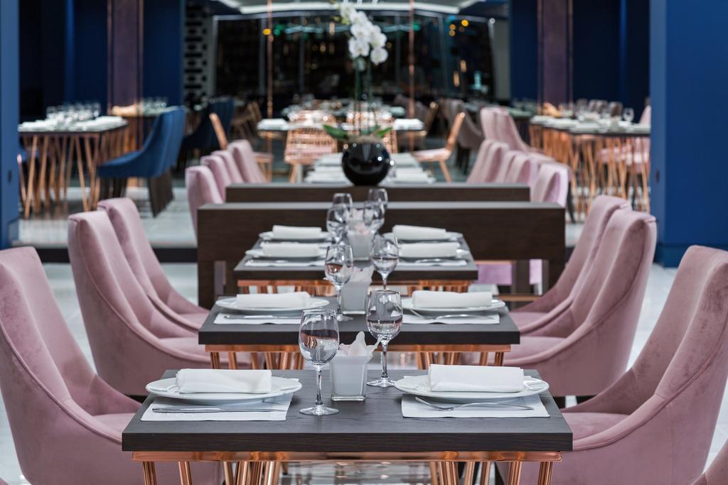 Sofitel Athens Airport Hotel Spata  Bagian luar foto The photo showcases a modern restaurant interior. In the foreground, there are several tables set with white plates, glassware, and napkins, arranged neatly. Each table has elegant pink chairs, giving a chic and contemporary vibe. In the background, 