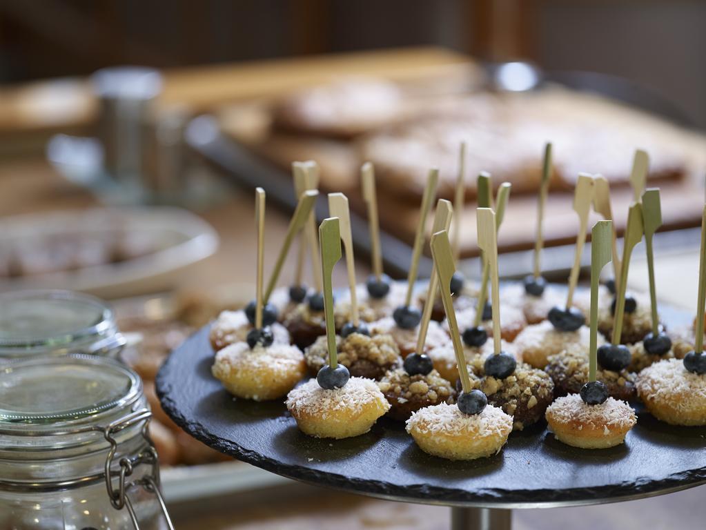 Sofitel Athens Airport Hotel Spata  Bagian luar foto The photo displays a dessert selection featuring small pastries arranged on a slate serving platter. The pastries are topped with coconut and have a single blue berry on each, with decorative sticks inserted for easy handling. In the background, ther