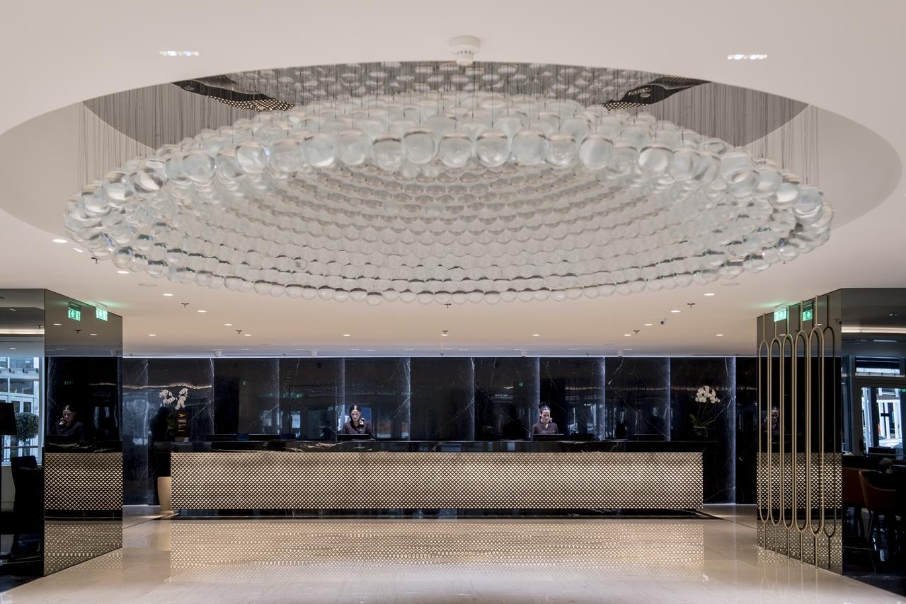 Sofitel Athens Airport Hotel Spata  Bagian luar foto The photo shows a modern hotel lobby. The centerpiece is an impressive, artistic chandelier made of glass, which hangs from the ceiling and features a circular design. Below the chandelier, there is a reception desk, which is sleek and stylish, likel