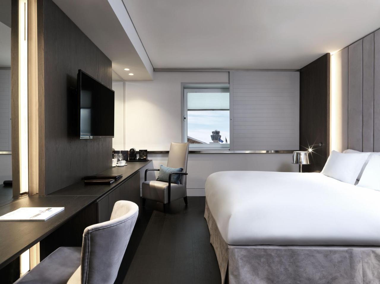 Sofitel Athens Airport Hotel Spata  Bagian luar foto The photo shows a modern hotel room. It features a large bed with white bedding, a sleek desk with a chair, and a flat-screen TV mounted on the wall. There are large windows allowing natural light to enter, and the room is decorated in neutral tones 